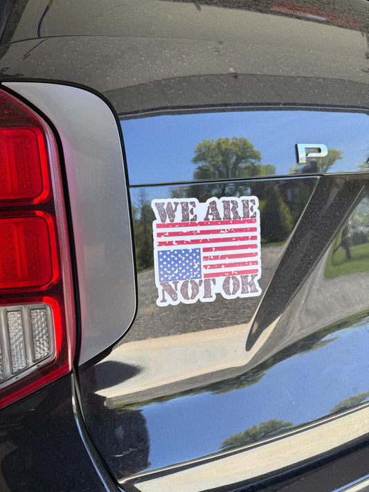 A vinyl decal or magnet featuring the phrase "We are not OK" in bold lettering, displayed alongside or over an upside-down American flag. The design symbolizes national distress and conveys a message of concern, protest, and social or political unrest.