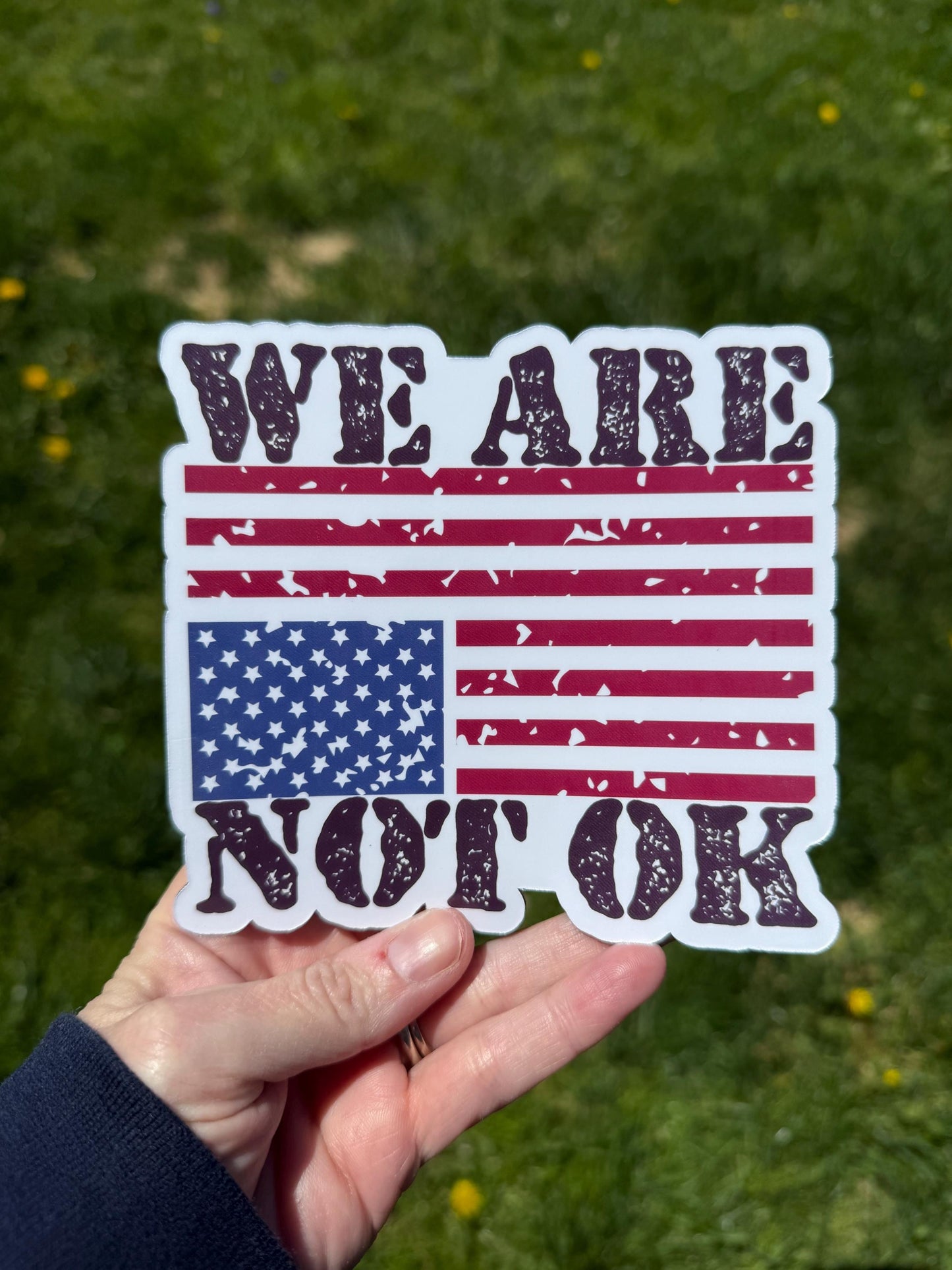 A vinyl decal or magnet featuring the phrase "We are not OK" in bold lettering, displayed alongside or over an upside-down American flag. The design symbolizes national distress and conveys a message of concern, protest, and social or political unrest.