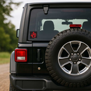A close-up rear view of a black off-road vehicle with a spare tire mounted on the back door. The vehicle is outdoors on a dirt road with blurred green trees in the background.