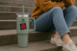 A sage green Stanley tumbler with a handle and clear lid sits on concrete steps next to a person wearing a mustard yellow sweater, blue jeans, and white sneakers.