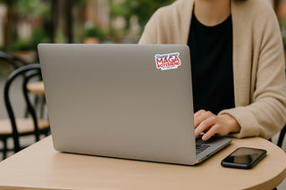 Person using a laptop with a dump your maga boyfriend sticker on the screen at an outdoor cafe.
