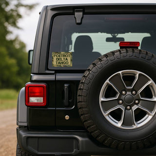 A black Jeep Wrangler rear view with a yellow 'FOXTROT DELTA TANGO' vinyl sticker on the back window, set against a blurred outdoor background.
