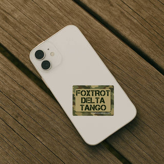 A white smartphone lying diagonally on a weathered wooden surface, with a rectangular camouflage patch on the back that reads “FOXTROT DELTA TANGO” in black letters.