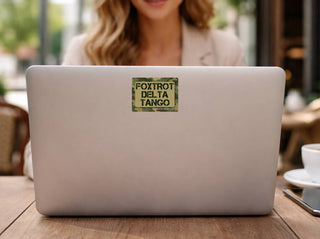 A square camouflage sticker with the text 'FOXTROT DELTA TANGO' in black capital letters, applied to the center of a silver laptop lid, shown in a cafe setting with a woman blurred in the background.