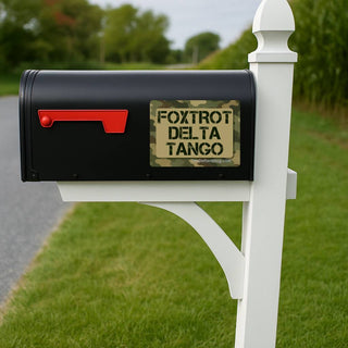 A black mailbox mounted on a white post in a grassy outdoor setting. The mailbox has a red flag and a decorative front label that reads “FOXTROT DELTA TANGO” with a small “Spartan Army Surplus” mark below it.