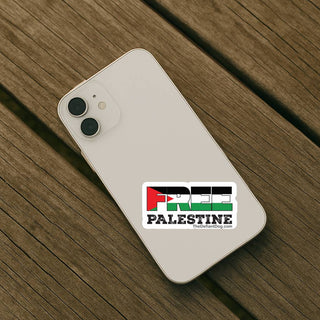A white smartphone lying diagonally on a weathered wooden surface, with a rectangular sticker on the back that reads “FREE PALESTINE” in red, green, black, and white flag colors. The phone brand is not visible, and the sticker text includes a small website credit.