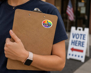 A person in a navy blue t-shirt holds a brown cardboard clipboard with a round rainbow-colored 'I VOTED' sticker on it. The background shows a blurred 'VOTE HERE' sign and an American flag.