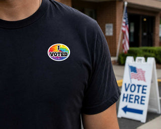 A close-up of a person wearing a black t-shirt with a circular 'I VOTED' pride sticker on the chest. The background shows a brick building, an American flag, and a white 'VOTE HERE' sign.