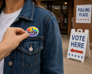 A person wearing a blue denim jacket has another person's hand pinning on a round rainbow-colored 'I VOTED' sticker. The background shows a polling place entrance with a 'VOTE HERE' sign.