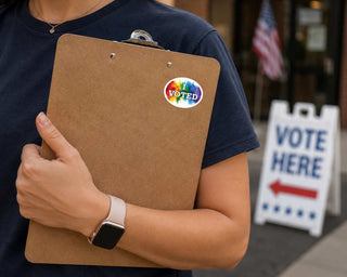 A person in a navy blue shirt holds a brown clipboard with a colorful oval sticker that says 'VOTED'. In the background, an American flag and a white 'VOTE HERE' sign are visible.