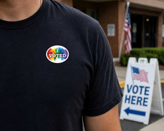 A close-up of a person wearing a black t-shirt with a circular 'I VOTED' graphic on the chest. The graphic features a vibrant rainbow watercolor background with white text.