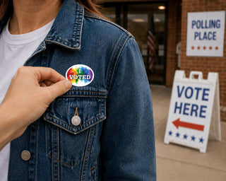 A person wearing a blue denim jacket has another person's hand pin a round sticker to the chest. The sticker features a rainbow design with the word 'VOTED' in white.