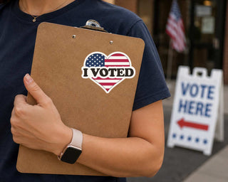 A person in a navy blue t-shirt holds a brown clipboard with a heart-shaped 'I VOTED' sticker featuring the American flag. In the background, a white 'VOTE HERE' sign is visible on a sidewalk.