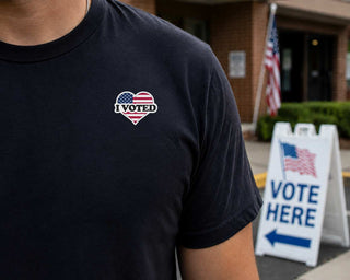 A close-up of a person wearing a black t-shirt with a heart-shaped graphic on the chest. The graphic features the American flag pattern with the text 'I VOTED' in white.