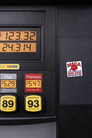 Funny anti-Trump MAGA Daddy Issues sticker displayed on the dark side panel of a gasoline pump. White square political decal with red MAGA lettering and a red baseball cap, photographed beside gas prices and fuel selection buttons.