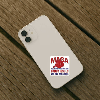 White smartphone resting on a rustic wooden table with a square MAGA Daddy Issues sticker attached to the back. Funny anti-MAGA political decal features a red hat illustration and bold red and navy blue lettering.