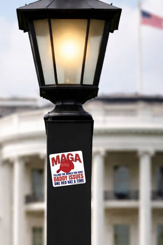 Anti-MAGA Daddy Issues political sticker displayed on a black street lamp post beneath a glowing lantern, with a softly blurred government-style building and American flag in the background. Funny anti-Trump red hat protest decal.