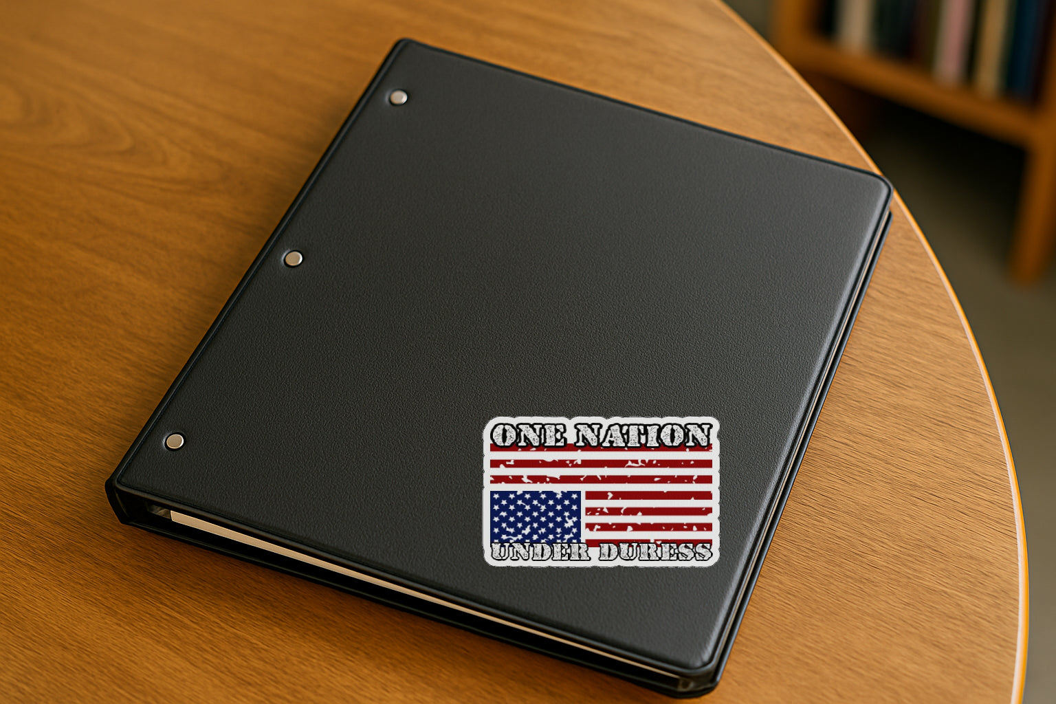 One Nation Under Duress' magnet or vinyl decal displayed on a black binder in a library. A bold, high-quality political sticker perfect for cars, laptops, fridges, or toolboxes. Great for activists, protestors, or anyone fed up with fascism.