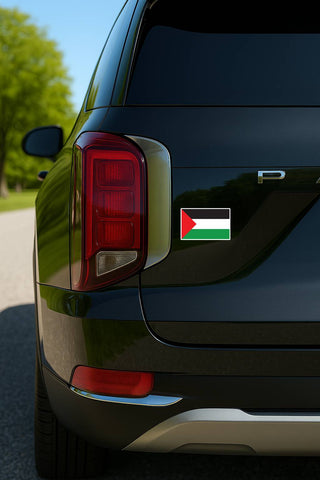 A small rectangular Palestinian flag decal sticker applied to the rear quarter panel of a black SUV, shown outdoors on a paved road with trees in the background.