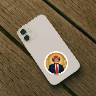 Round political satire sticker featuring a clown-style Trump portrait with rainbow hair and halo applied to the back of a smartphone, demonstrating use on phones and personal devices.