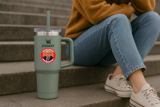 A sage green Stanley tumbler with a handle and clear lid sits on concrete steps next to a person sitting on the same steps. The tumbler has a circular sticker featuring a portrait of Donald Trump with the text 'TWORLD'S BIGGEST EMBARRASSMENT'.