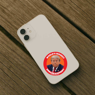 A white smartphone lying on a weathered wooden surface, with a circular red sticker graphic on the back featuring a portrait of Donald Trump and the text 'WORLD'S BIGGEST EMBARRASSMENT'.