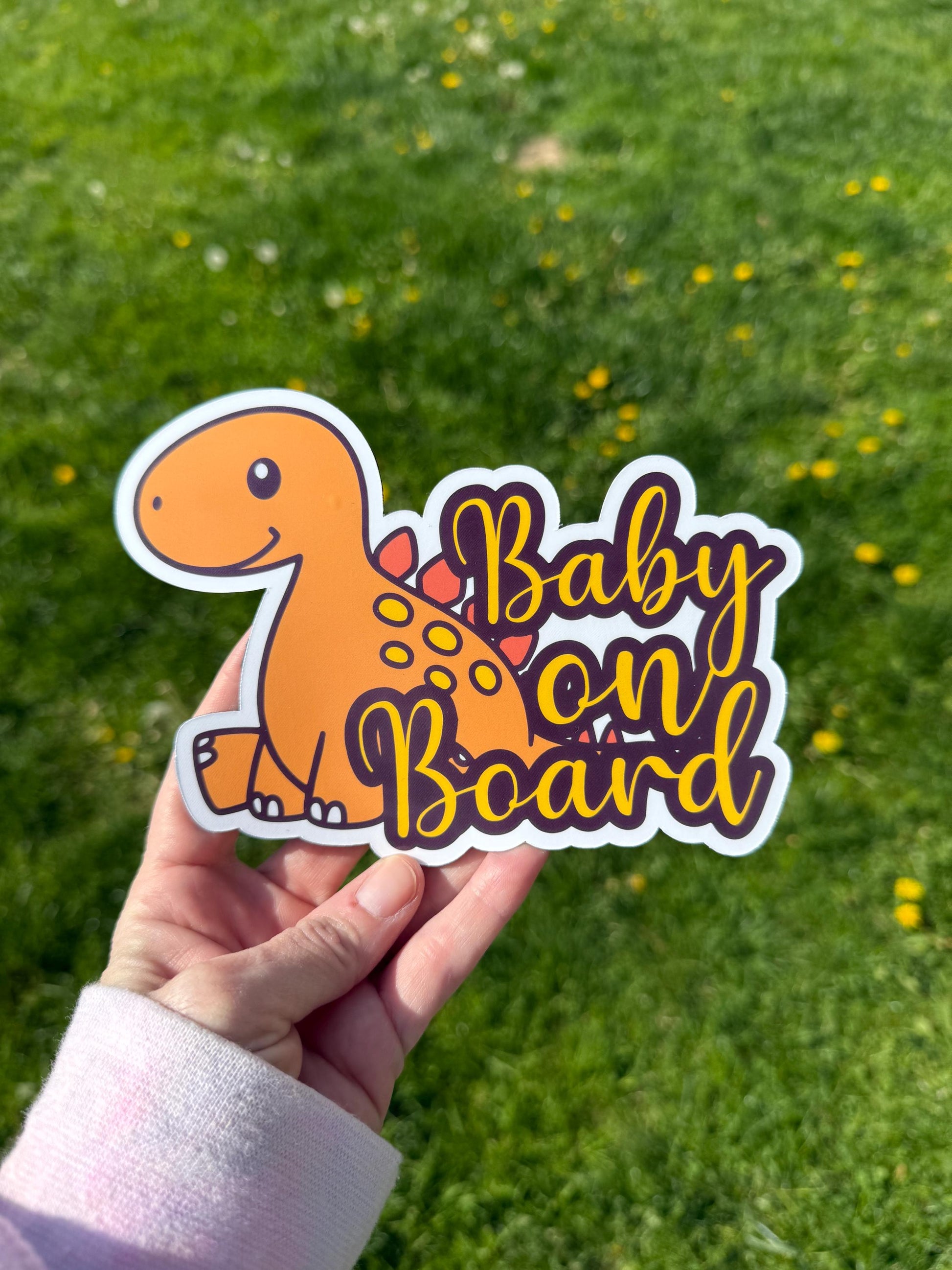 “Baby on Board” orange dino decal held in hand above green grass and dandelions.