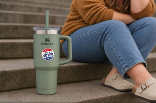 Round “Be a Voter” sticker on a sage green Stanley tumbler beside a person sitting on outdoor steps.