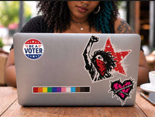 “Be a Voter” sticker on a silver laptop alongside colorful statement stickers at a café table.