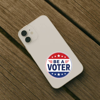 Round red, white, and blue “Be a Voter” sticker on the back of a white iPhone resting on a wooden table.