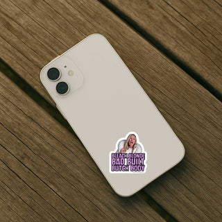 Light colored iPhone resting on wooden surface with Bleach Blonde Bad Built Butch Body sticker applied to the back. Compact vinyl decal perfect for phone cases and small accessories.
