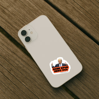 A white smartphone lying on a weathered wooden surface, with a die-cut sticker on the lower back showing a portrait and the text 'BONE SPUR HERO 1969' and 'TheDailyJester.com'.