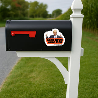 A black mailbox mounted on a white post outdoors, with a red flag and a die-cut sticker on the side showing a portrait and the text 'BONE SPUR HERO 1968'.