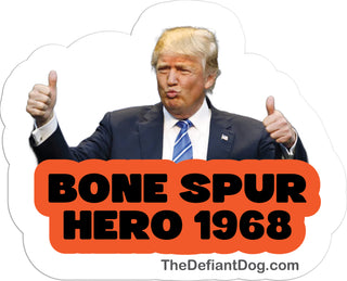 A die-cut sticker featuring a photo of Donald Trump giving two thumbs up, with the text 'BONE SPUR HERO 1968' in black letters inside an orange cloud shape. The sticker is shown on a white background with the website 'TheDefiantDog.