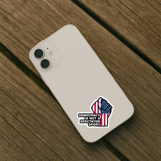 Light colored iPhone with Democracy Is Not a Spectator Sport protest sticker applied to the back. Small waterproof vinyl decal with American flag raised fist design.