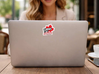A close-up of a silver laptop on a wooden table, featuring a red and white 'Elbows Up of Canada' sticker on the lid. A woman is visible in the blurred background, and a white coffee cup sits on the right.