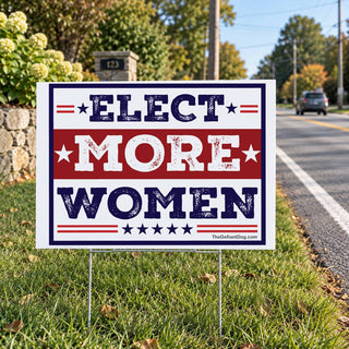 Elect More Women election lawn sign displayed beside a residential road, featuring distressed navy blue and red lettering with stars and stripes.
