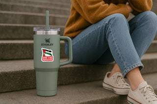 A sage green Stanley travel tumbler with a handle and lid, featuring a custom sticker of the Palestinian flag and the text 'I LOVE ISRAEL', placed on concrete steps next to a person sitting on stairs wearing a mustard sweater and blue jeans.