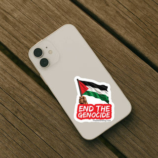 A white smartphone lying on a wooden surface, showing a die-cut sticker on the back. The sticker features the Palestinian flag, a raised hand, and the text 'END THE GENOCIDE' in white on a red background.