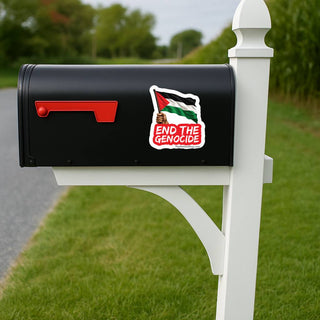 A black mailbox on a white post with a red flag, shown outdoors on a grassy lawn beside a road. A sticker on the side of the mailbox features the Palestinian flag and the text 'END THE GENOCIDE'.