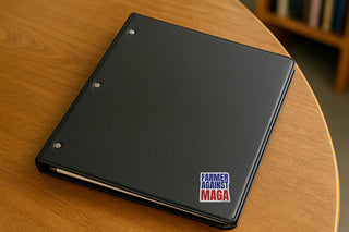 Farmer Against MAGA vinyl sticker attached to the corner of a black three-ring binder on a wooden table.