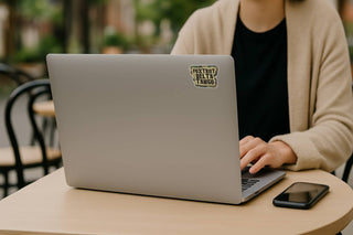 Person using a laptop with a sticker on the back, sitting at an outdoor table.