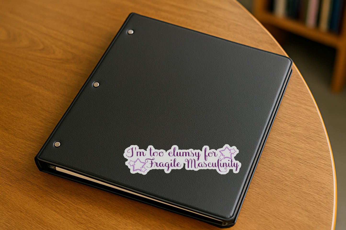 I'm Too Clumsy for Fragile Masculinity' magnet or vinyl decal displayed on a black binder in a library. A bold, high-quality political sticker perfect for cars, laptops, fridges, or toolboxes. Great for activists, protestors, or anyone fed up with fascism.