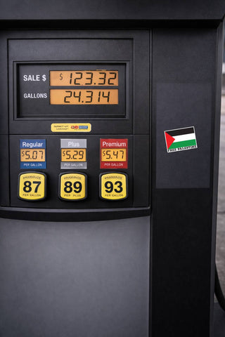 Close-up of a black fuel pump display showing sale amount, gallons, and three fuel options labeled Regular, Plus, and Premium, with a small Palestinian flag sticker on the side.