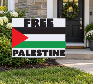 A rectangular yard sign on metal stakes in a front yard, featuring the Palestinian flag and the words “FREE PALESTINE” in bold black letters. The sign is photographed outdoors in front of a house entrance with grass, shrubs, and a door wreath visible.