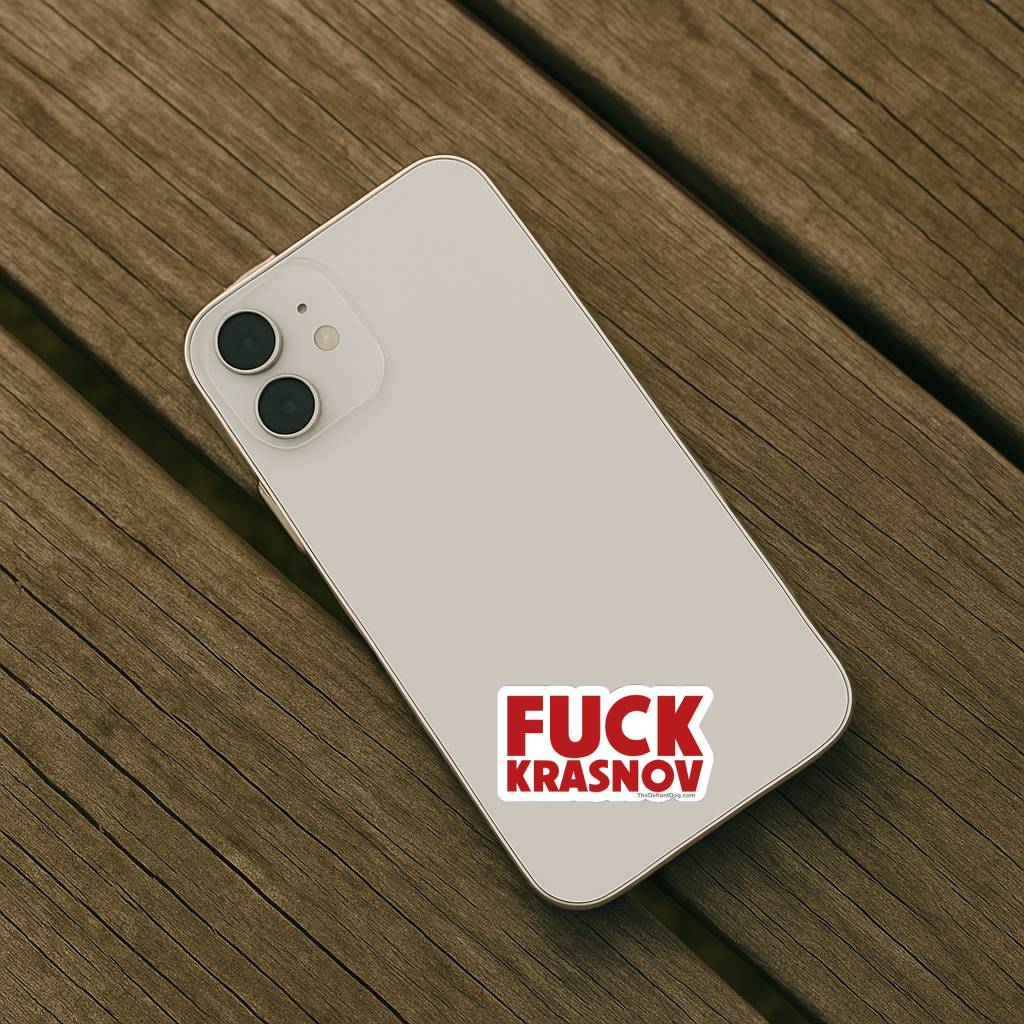 A light-colored smartphone placed on a wooden surface with the Fuck Krasnov sticker applied to the back. The compact size shows how the design works on smaller personal items.