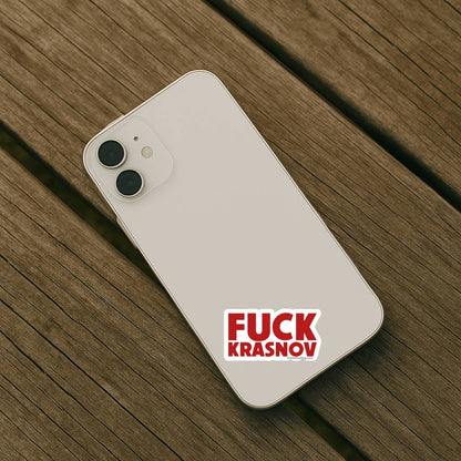 A light-colored smartphone placed on a wooden surface with the Fuck Krasnov sticker applied to the back. The compact size shows how the design works on smaller personal items.