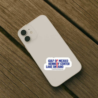 A white smartphone case shown from the back, placed on a wooden plank surface. The case has a printed sticker reading "GULF OF MEXICO KENNEDY CENTER LAKE ONTARIO" in blue and red text on a white cloud shape, with "TheDefiantDog.
