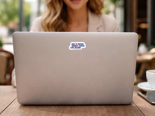 A close-up of a silver laptop on a wooden table, with a small white and blue sticker reading 'GULF OF MEXICO BETWEEN CENTER LAKE OR CANAL' on the lid. A woman is blurred in the background, and a coffee cup is visible on the right.