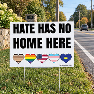 A white rectangular yard sign with the black text HATE HAS NO HOME HERE. Below the text are five hearts featuring the inclusive pride flag, rainbow flag, American flag, transgender flag, and a blue heart with a yellow equal sign.
